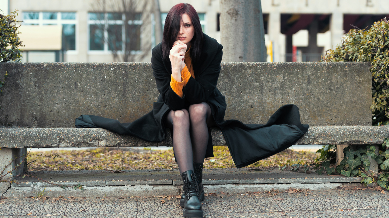 Девушка в черном пальто сидит на бетонной лавке 