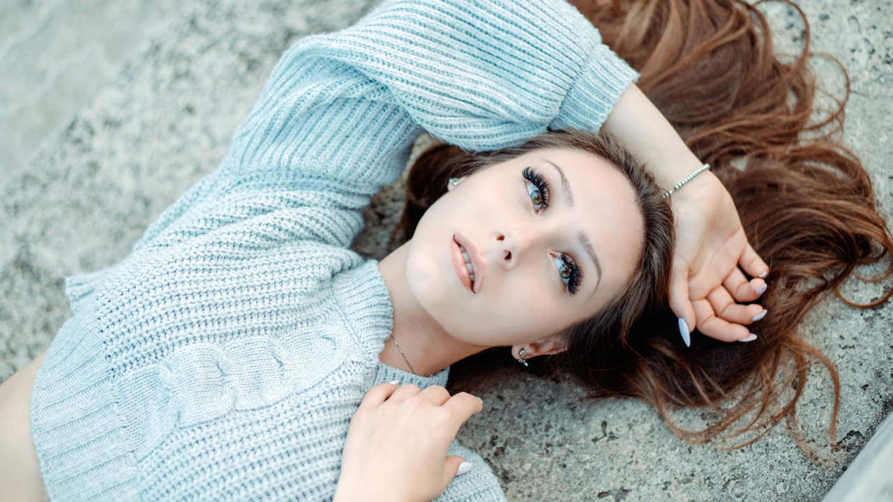 Длинноволосая девушка в голубом свитере лежит на земле 