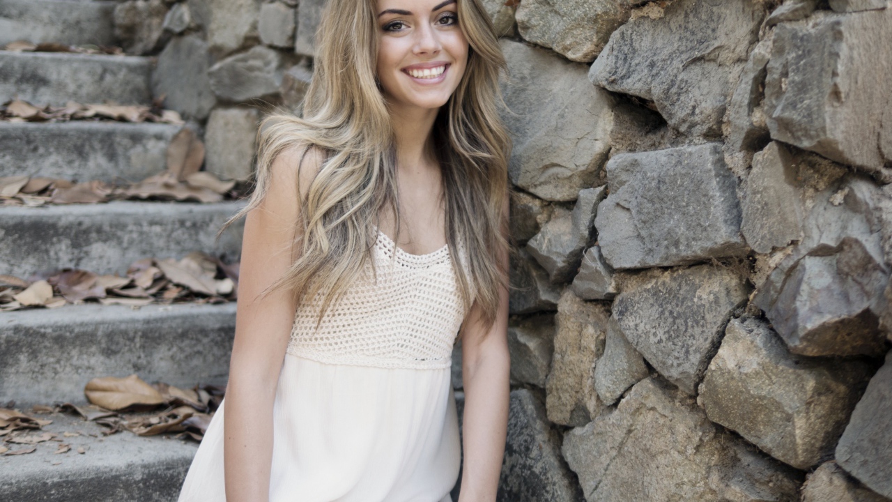 Улыбающаяся  девушка в белом платье стоит на ступеньках у стены