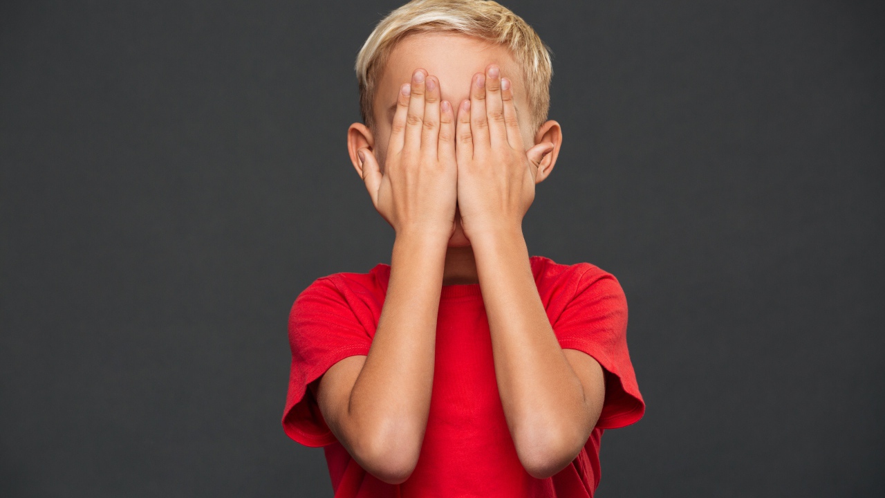 Маленький мальчик в красной футболке закрыл лицо руками 