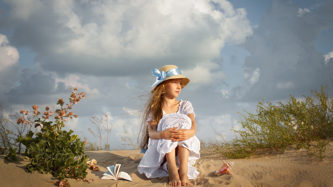 Маленькая девочка в белом платье сидит на песке 
