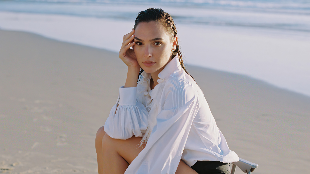 Актриса Галь Гадот в белой рубашке сидит на песке