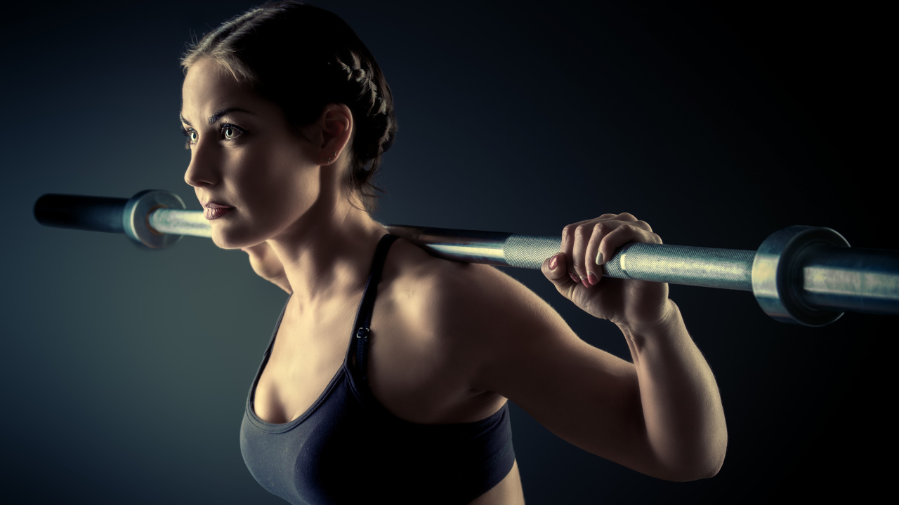 The sports girl is engaged with a barbell in the gym on a gray background