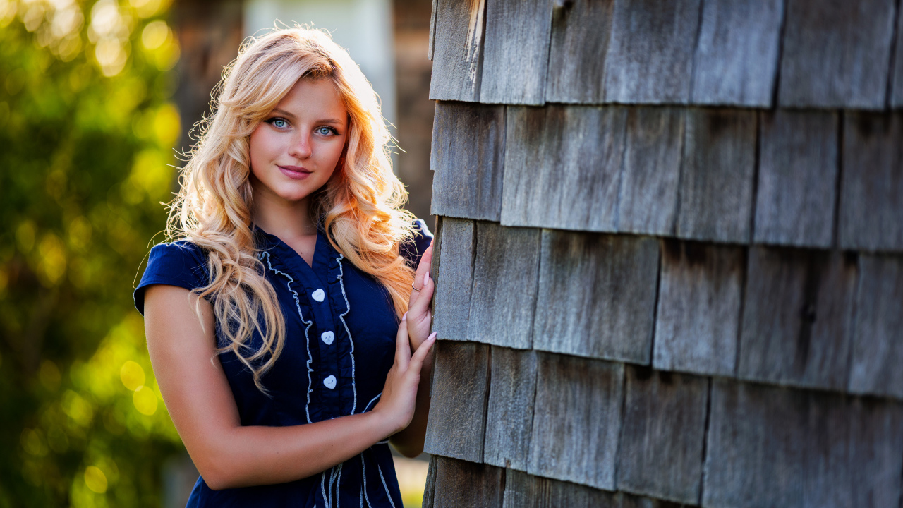 Голубоглазая блондинка в платье стоит у стены 