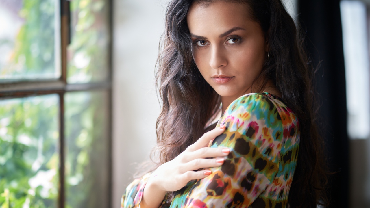 Длинноволосая кареглазая девушка стоит у окна