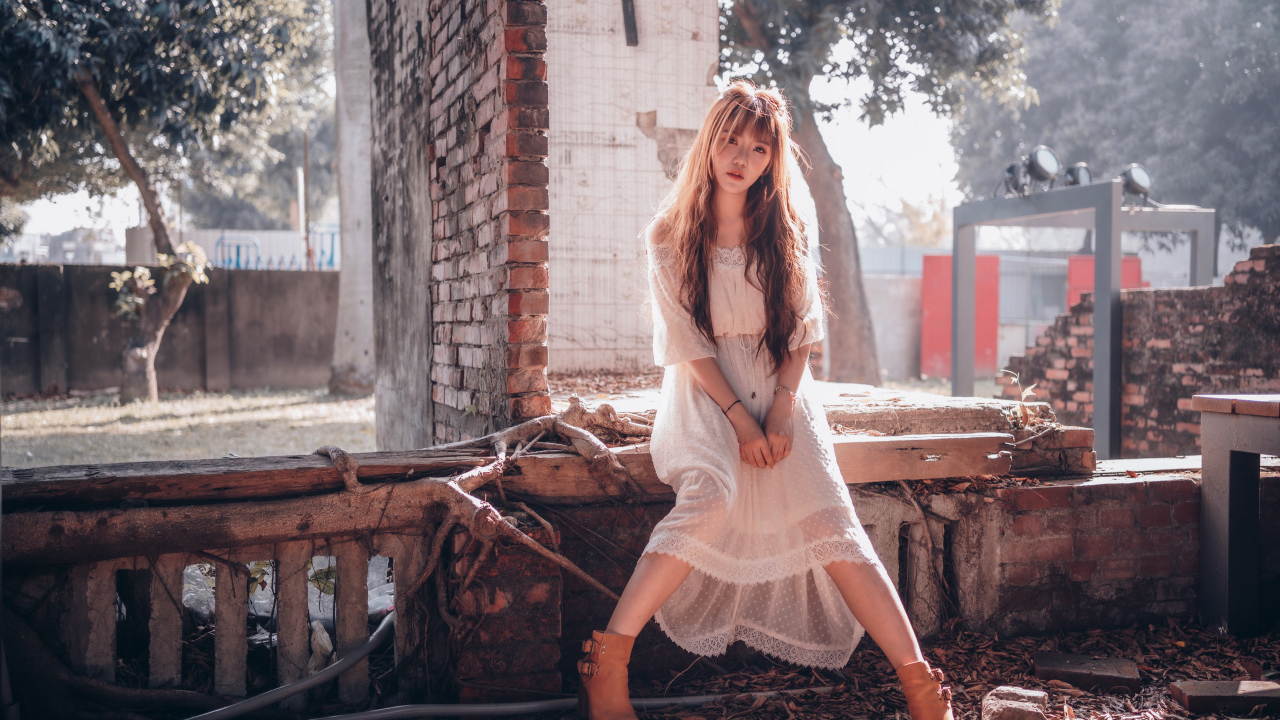 Молодая девушка азиатка в белом платье сидит на разваленной стене