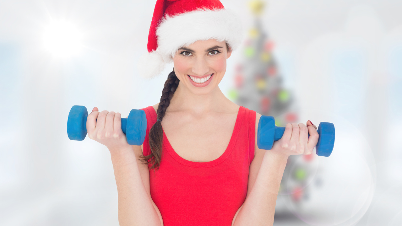 Красивая девушка в шапке Санта Клауса с гантелями в руках 