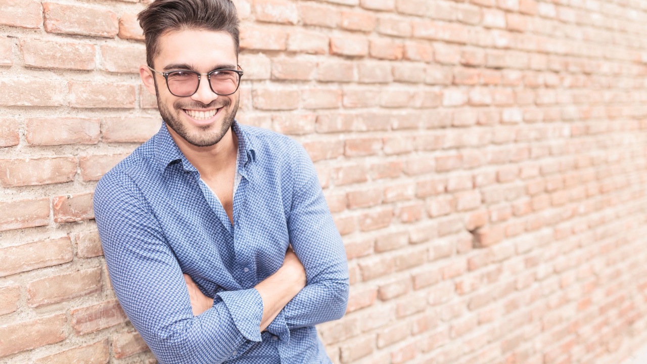 Улыбающийся мужчина в очках стоит у стены 