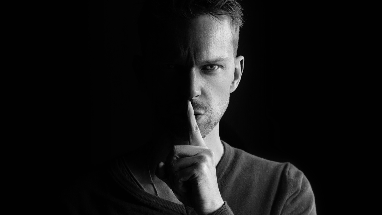 Молодой мужчина закрыл рот пальцем на черном фоне 