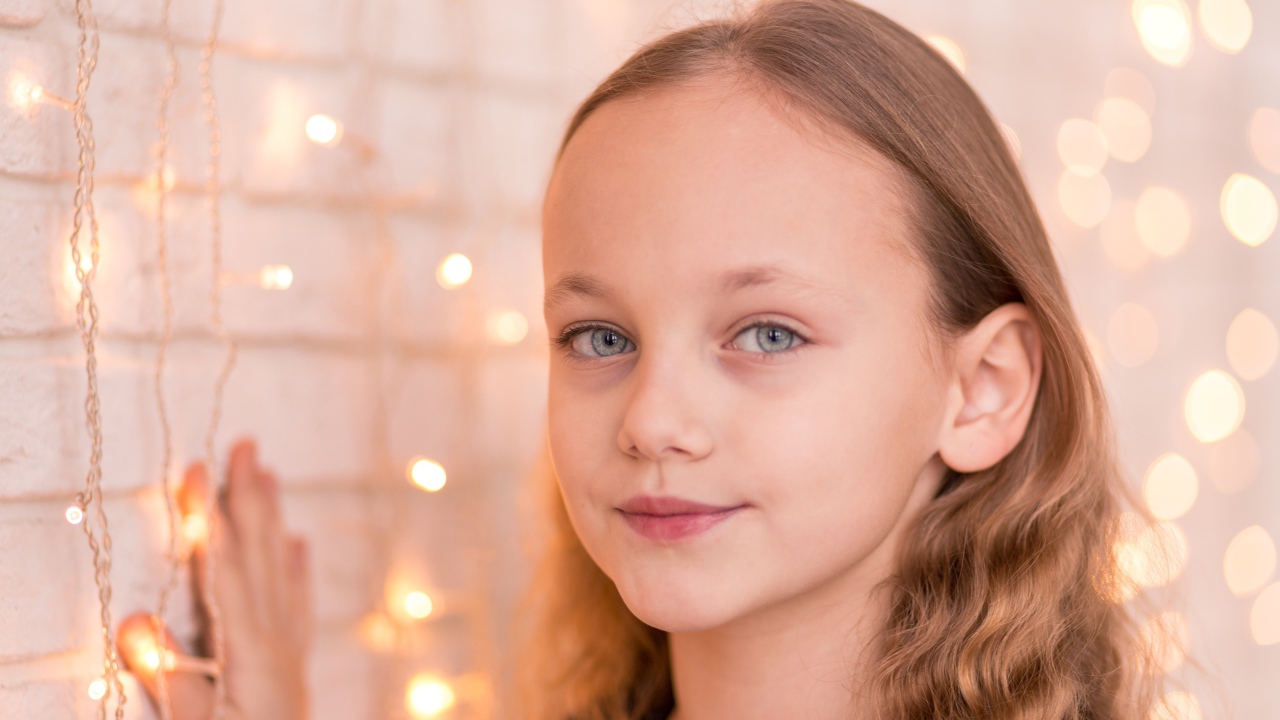 Красивая голубоглазая девочка стоит у стены с гирляндой