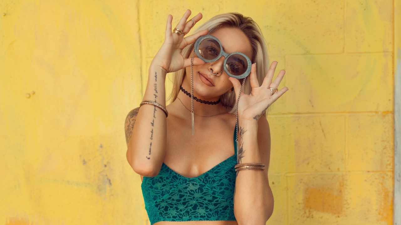 Девушка в очках с татуировками на руках стоит у стены