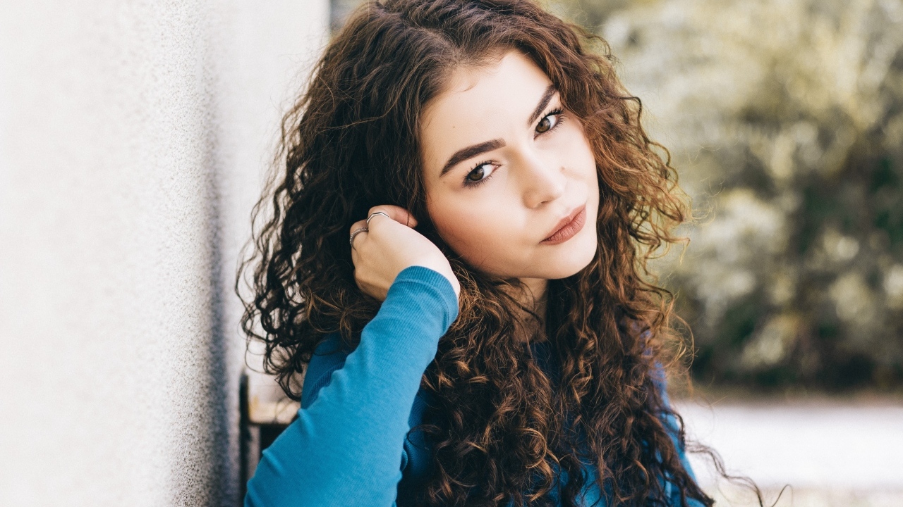 Кареглазая девушка с кудрявыми волосами стоит у стены