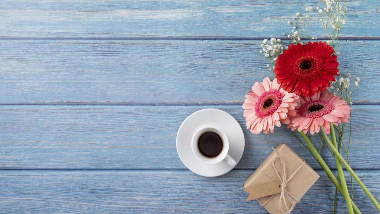 Три герберы с чашкой кофе и подарком на синем столе