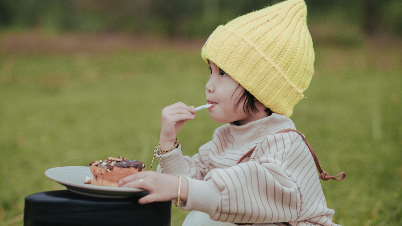 Маленькая девочка в желтой шапке ест пирожное