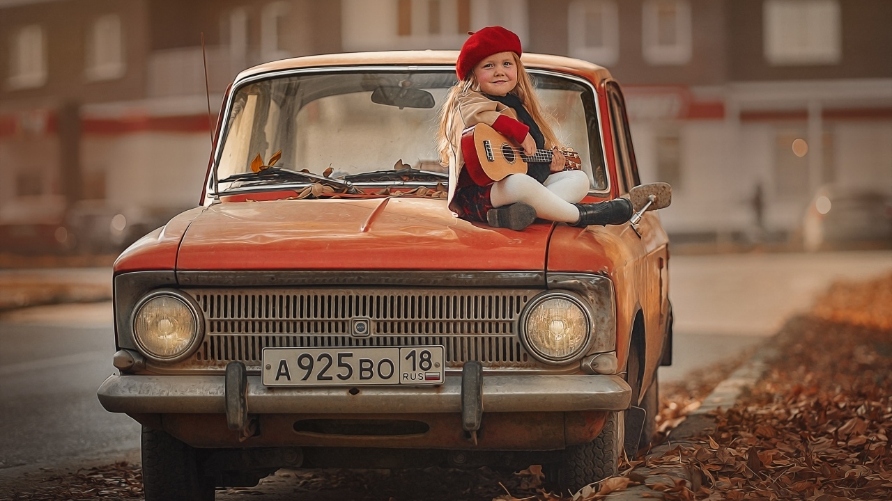 Маленькая девочка с гитарой сидит на красном автомобиле осенью