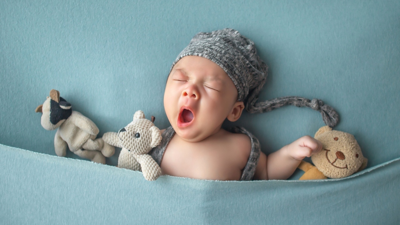 Зевающий малыш с игрушками под серым покрывалом