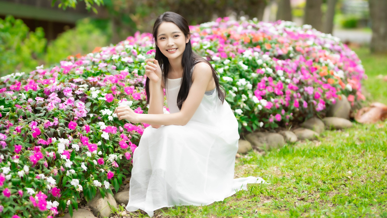 Милая улыбающаяся азиатка в белом платье сидит у цветов