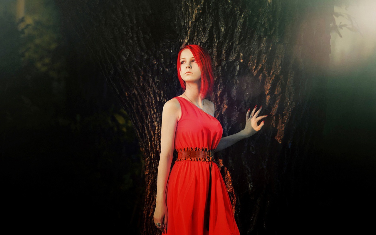 Рыжая девушка в красном платье стоит у дерева
