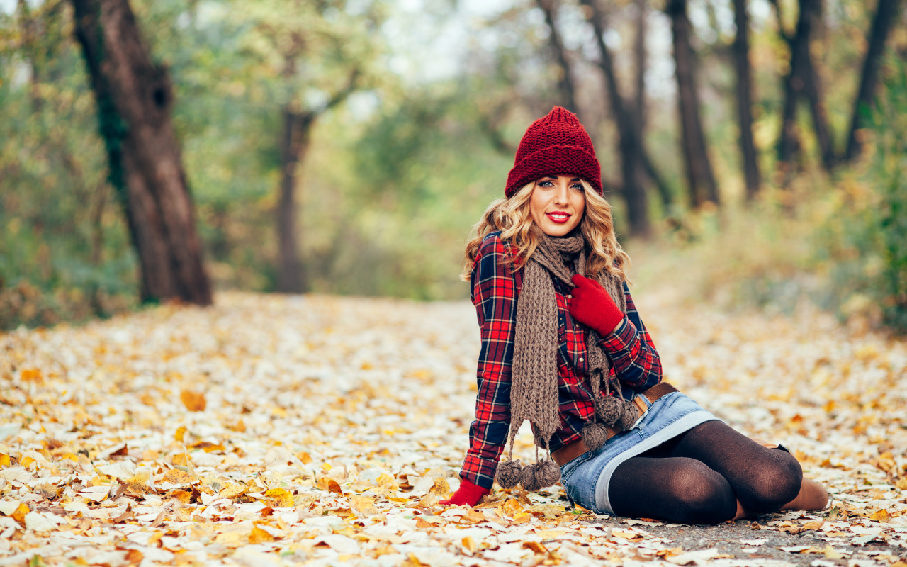 Молодая улыбающаяся девушка в красной шапке сидит на листве осенью