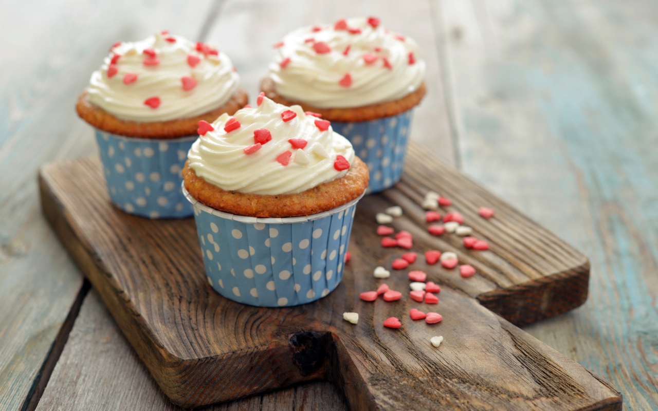 Три кекса со сливками украшены сердечками стоят на разделочной доске