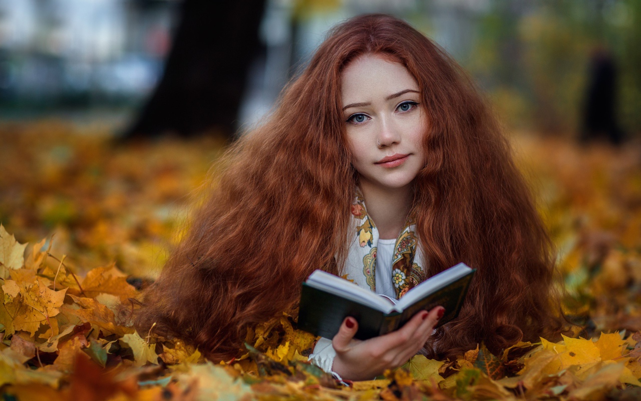 Рыжеволосая девушка с книгой лежит на опавших листьях осенью 