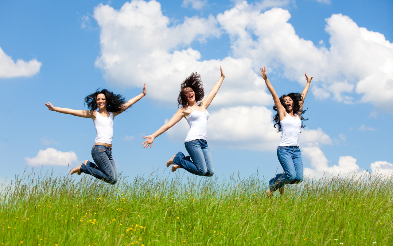 Три молодые девушки прыгают в зеленой траве на фоне неба 