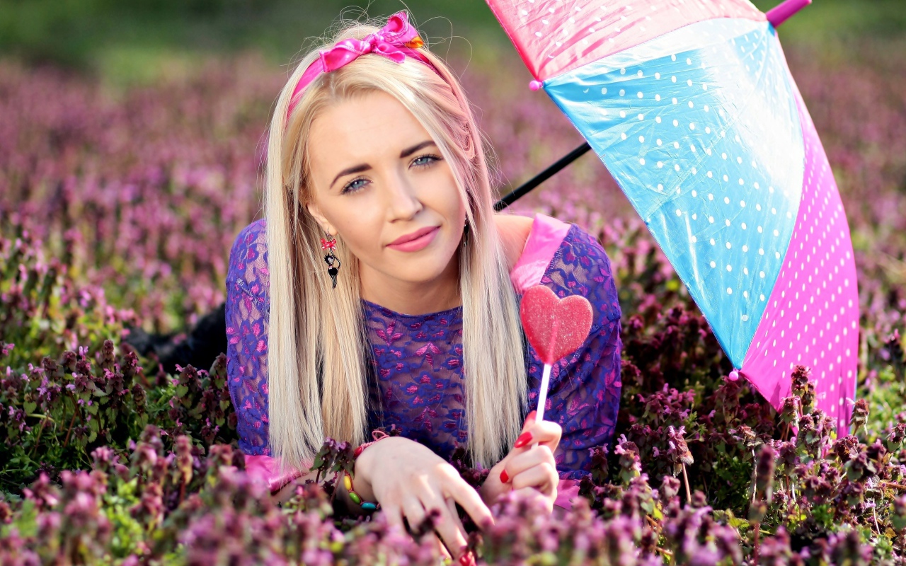 Молодая девушка блондинка лежит на поляне с сиреневыми цветами под разноцветным зонтом