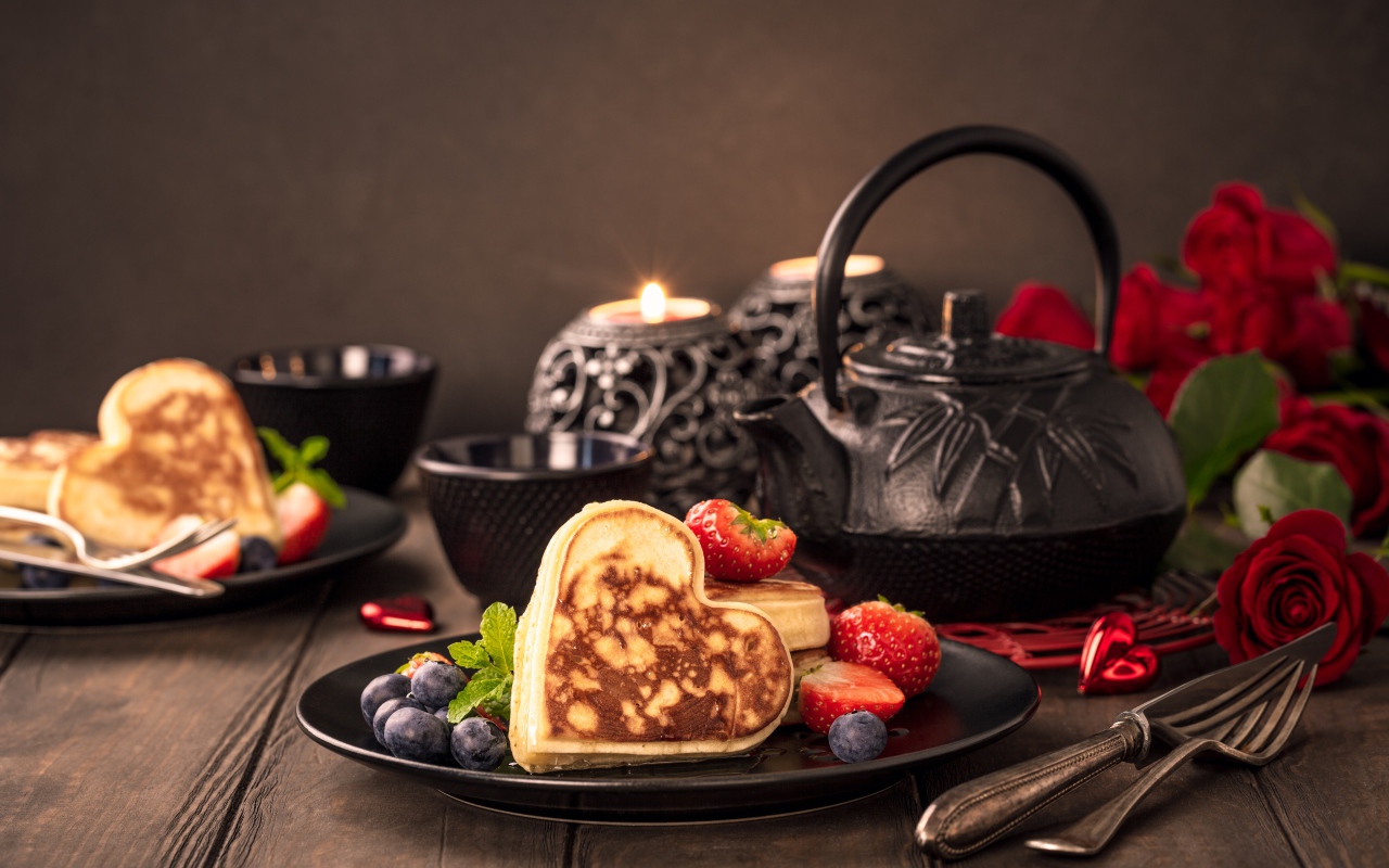 Оладьи в форме сердца с ягодами черники и клубники на столе с чаем и свечами