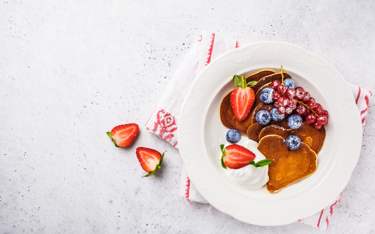 Оладьи со сметаной в тарелке с ягодами черники, клубники и красной смородины