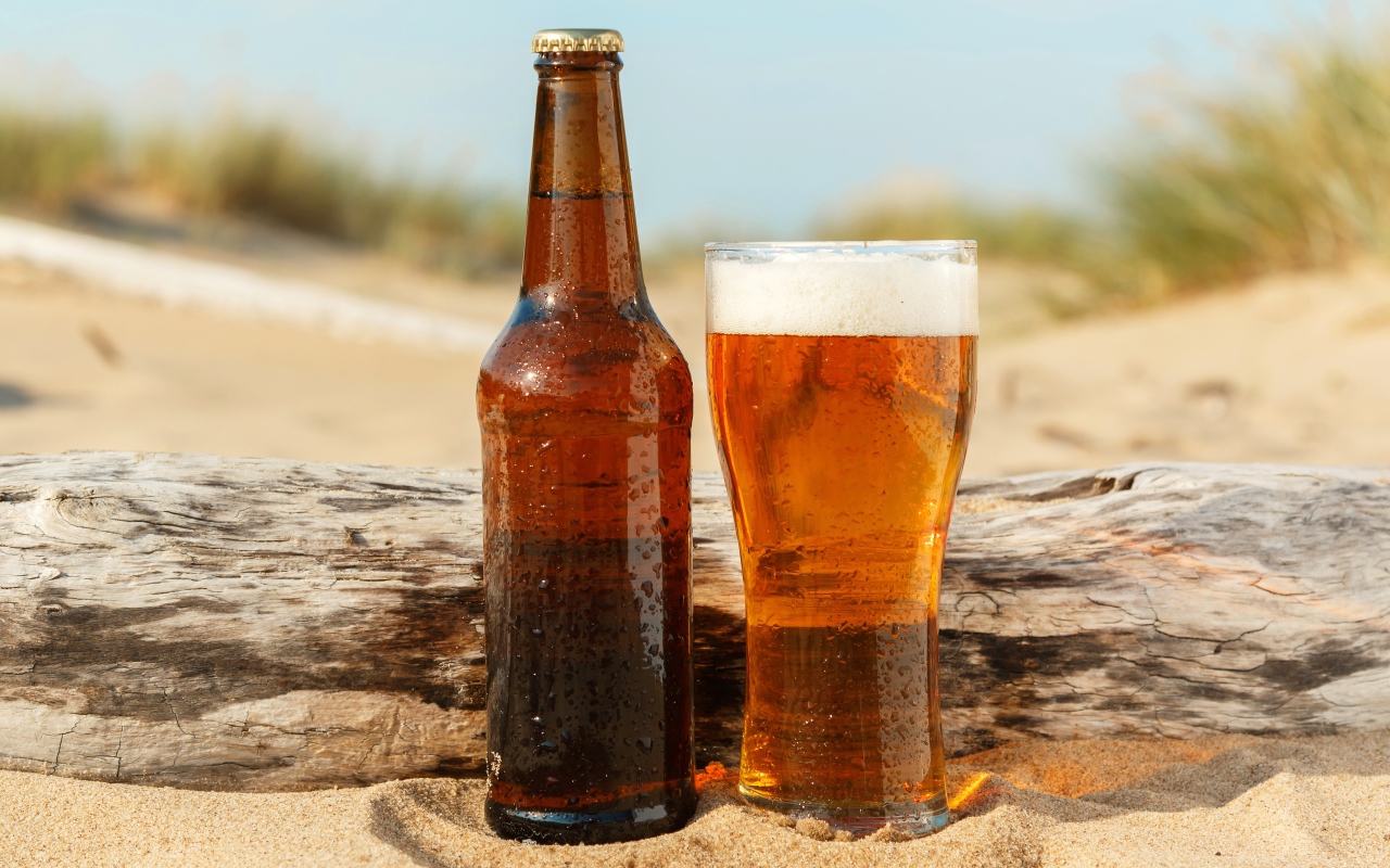 Бутылка пива и бокал стоят на песке 