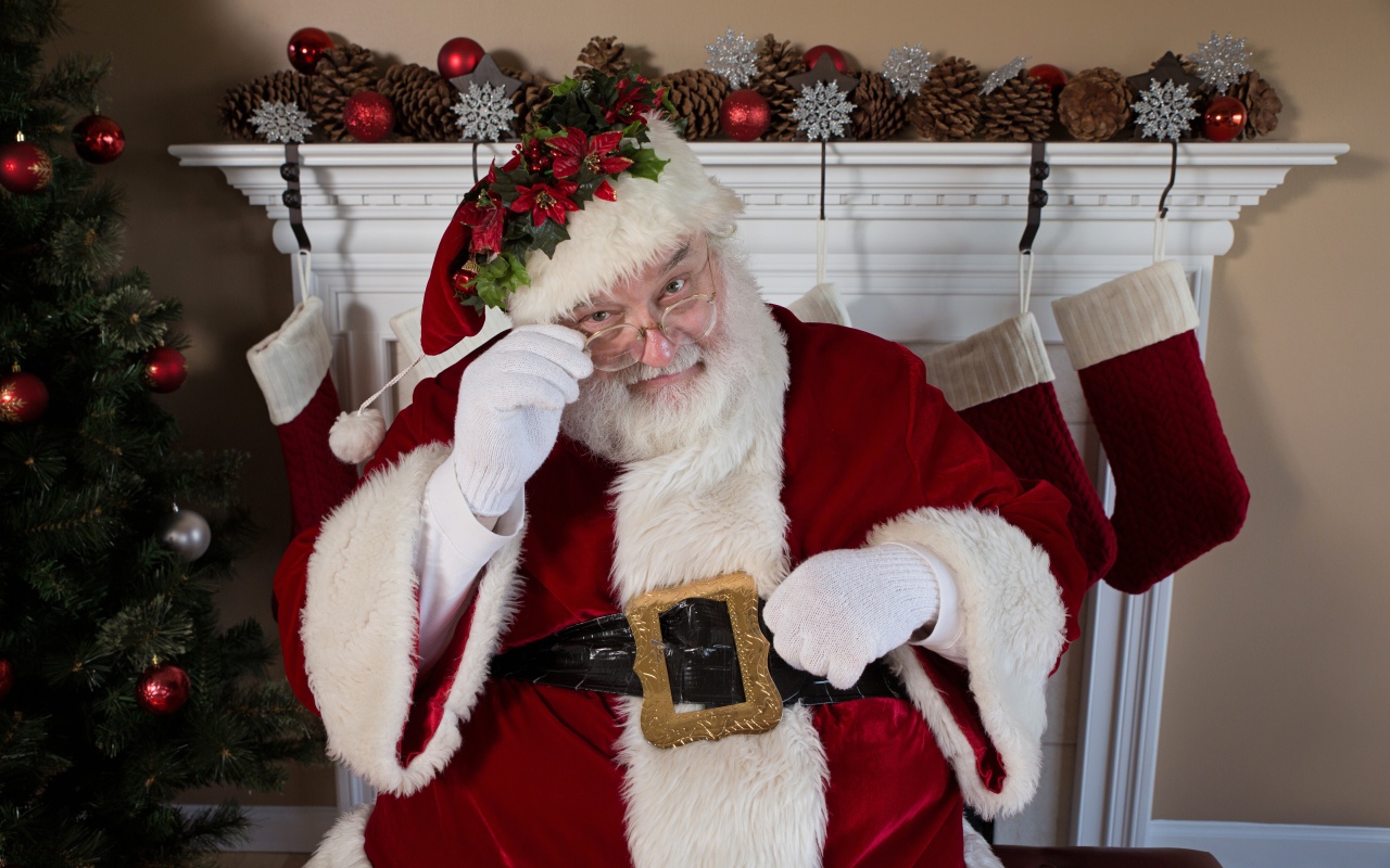 Санта Клаус в очках сидит у камина у новогодней елки