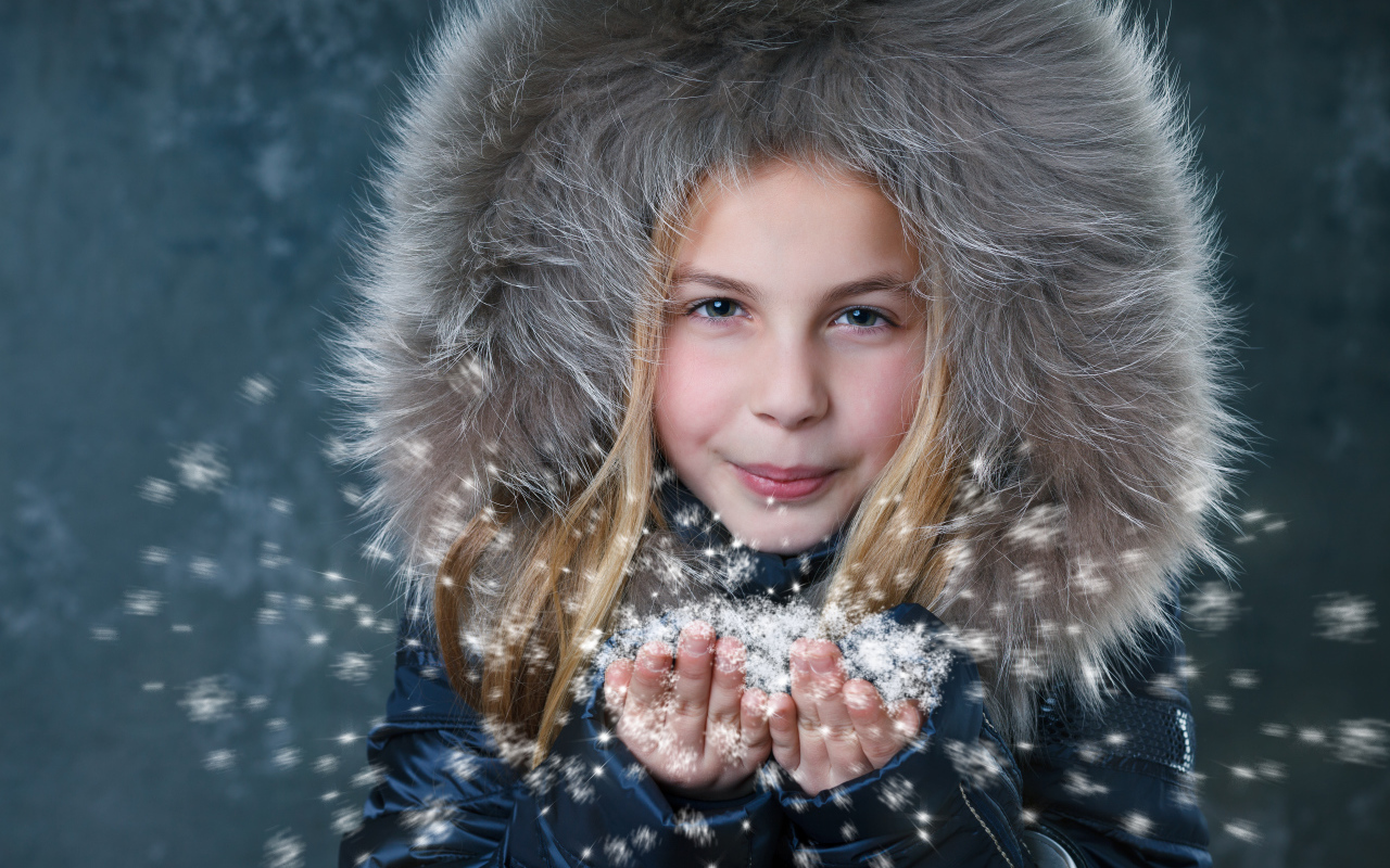 Маленькая девочка в капюшоне сдувает снежинки с ладоней