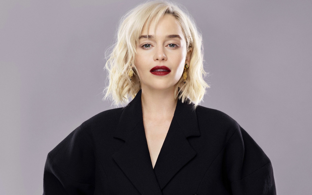 Яркая блондинка, актриса Эмилия Кларк в черном пальто на сером фоне