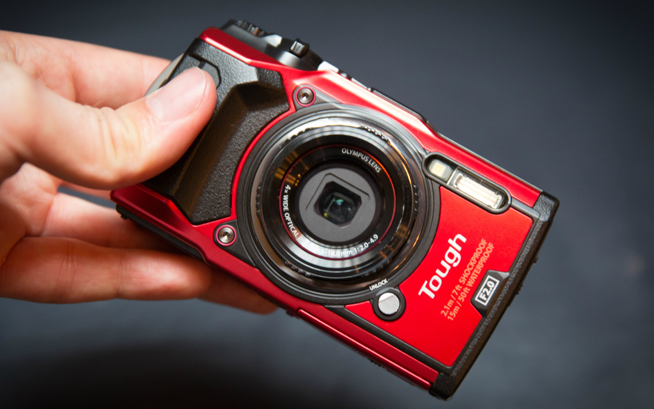 Красный фотоаппарат Olympus Tough TG-5 в руке на сером фоне