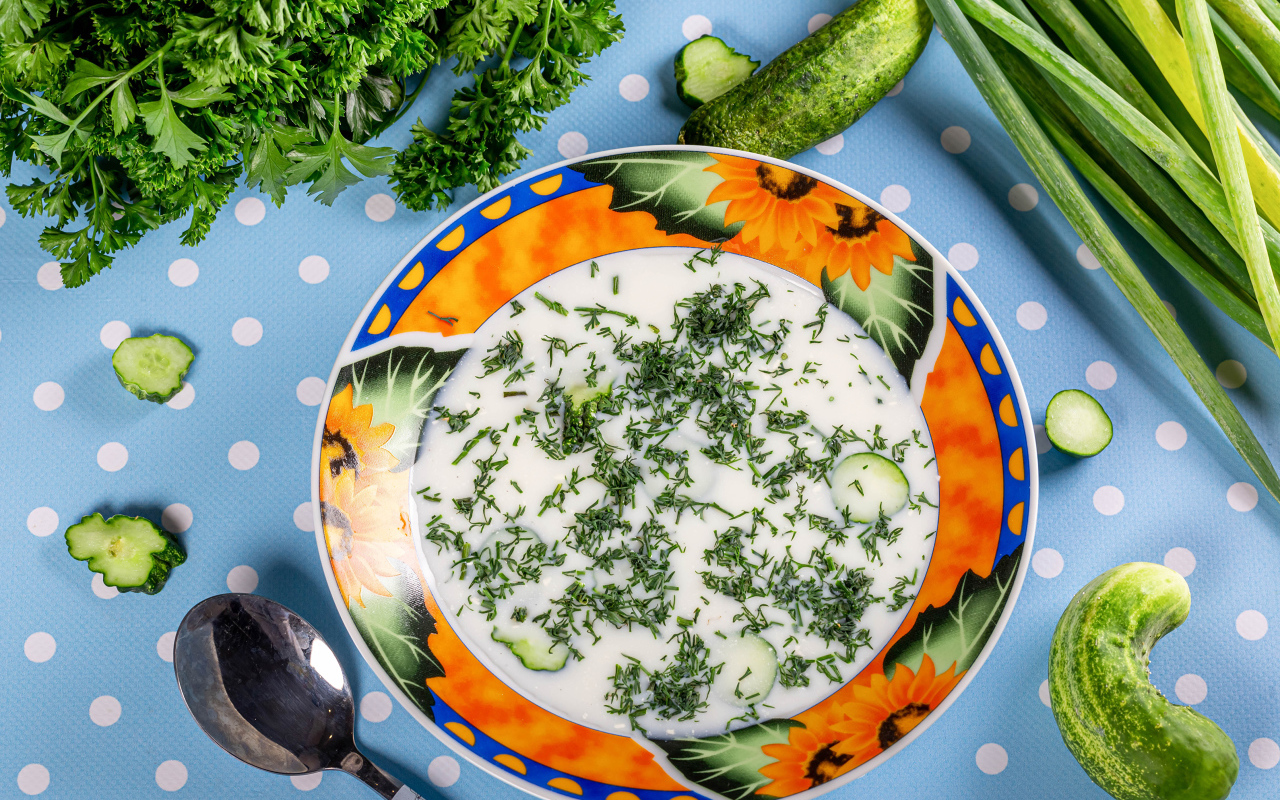 Окрошка в тарелке на столе с зеленью и огурцами