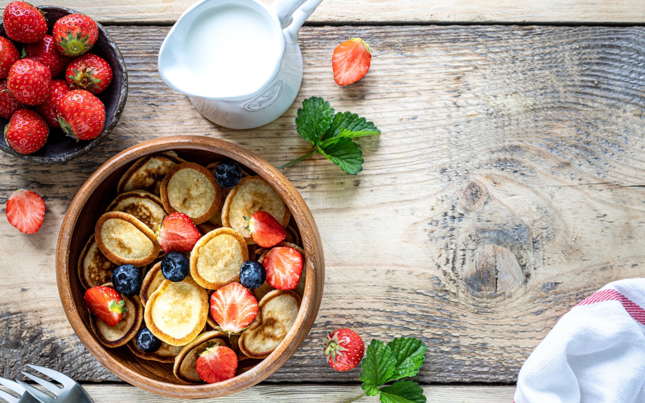 Маленькие оладьи в миске с клубникой и черникой на столе с молоком 