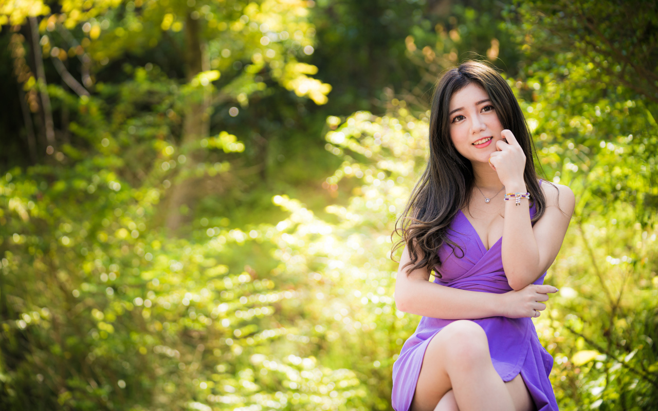 Улыбающаяся девушка азиатка в фиолетовом платье сидит в парке 