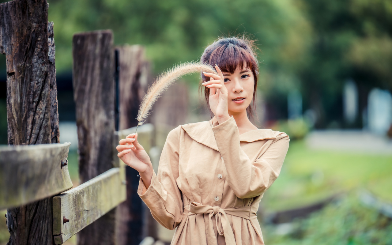 Девушка азиатка в пальто стоит у забора 