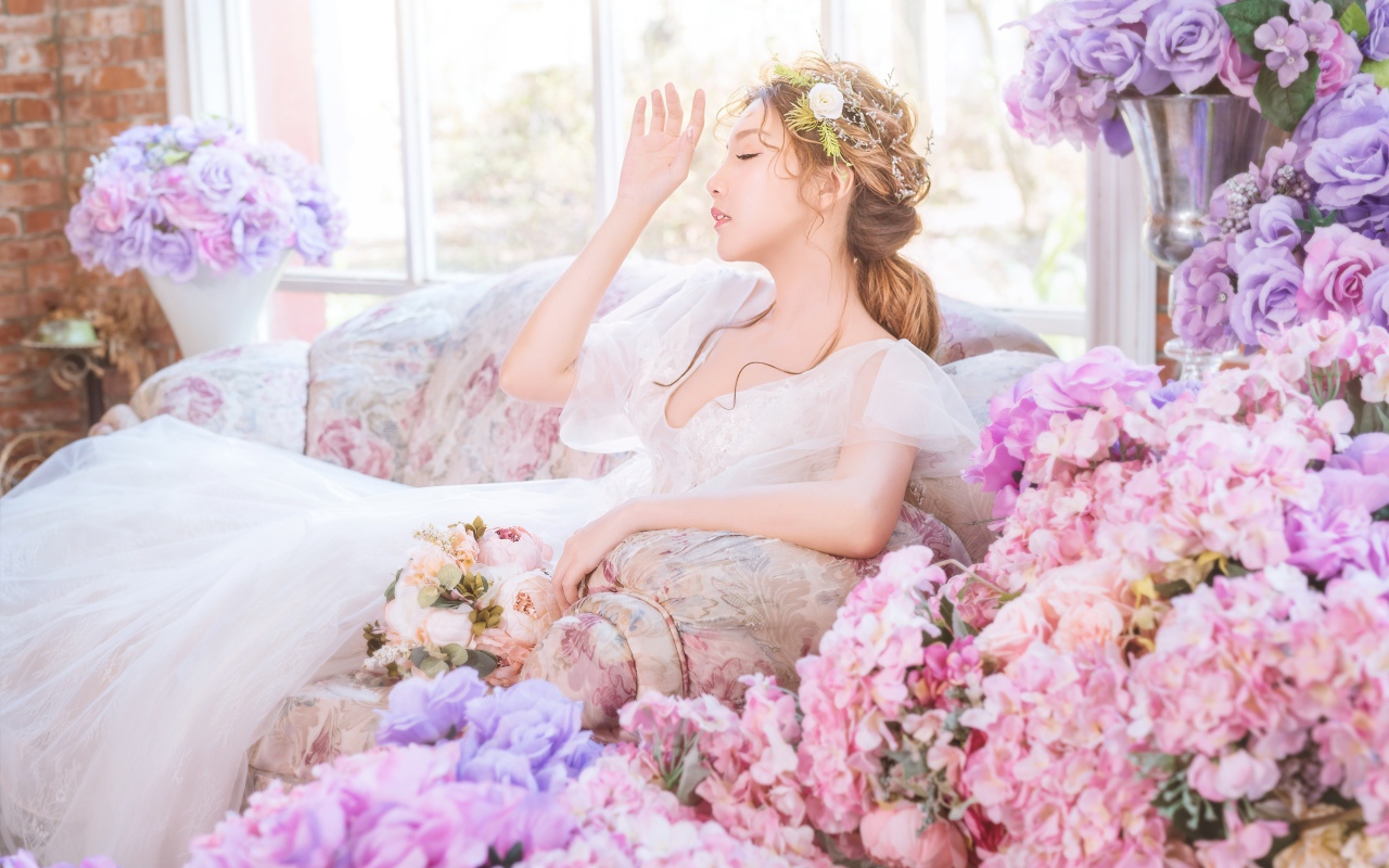 Девушка азиатка в белом платье сидит на диване в комнате с цветами