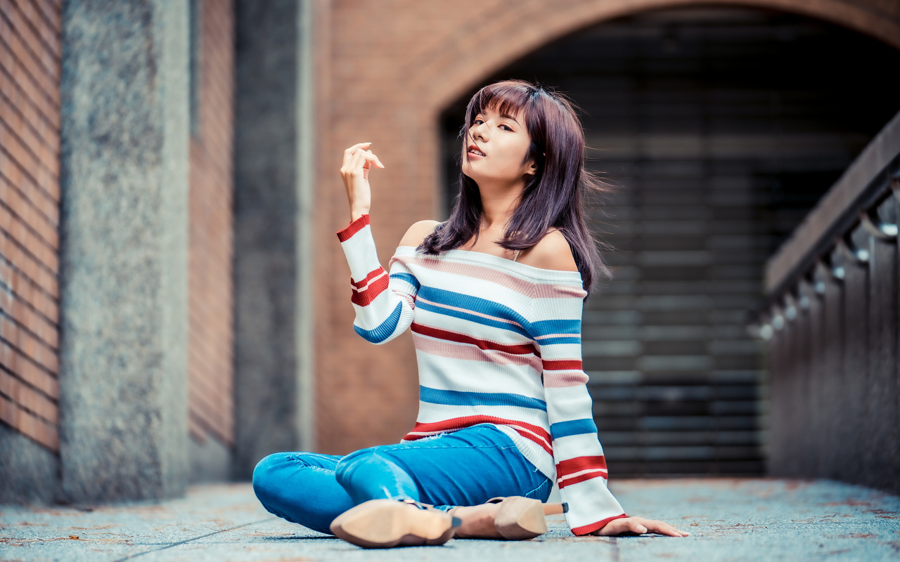 Девушка азиатка в джинсах и полосатом свитере сидит на асфальте