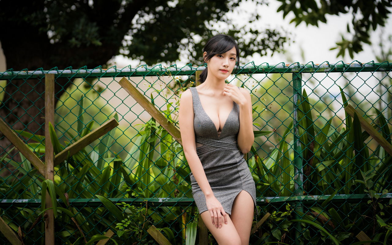 Красивая азиатка в сером платье стоит у зеленой сетки 