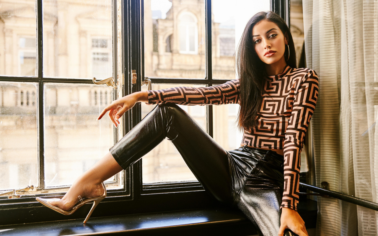 Красивая брюнетка в черных брюках сидит на окне 