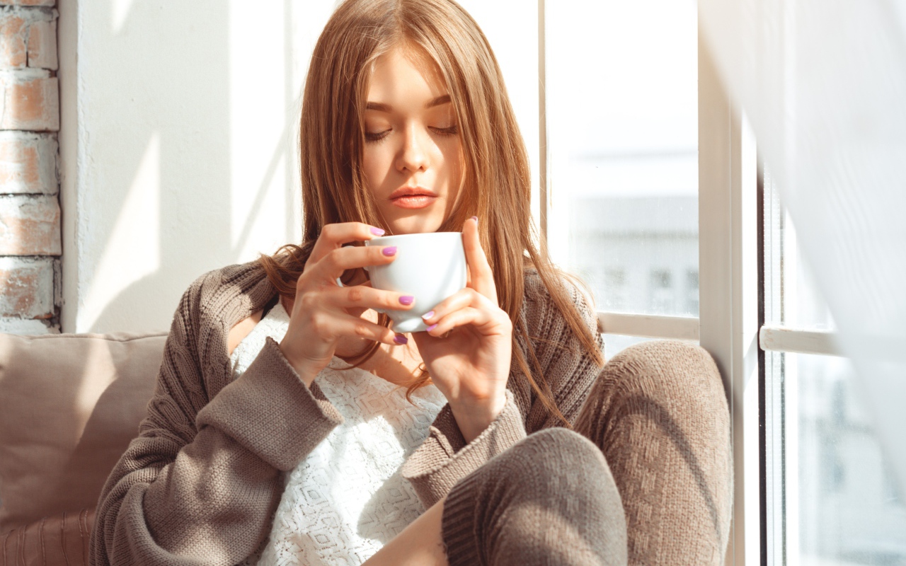 Красивая девушка в свитере пьет кофе у окна