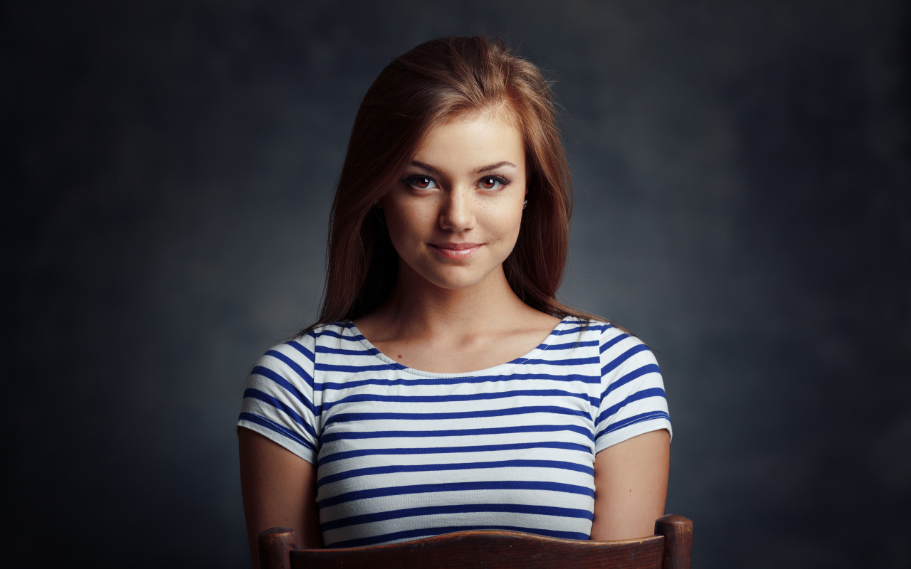 Милая кареглазая девушка в полосатой майке сидит на стуле 