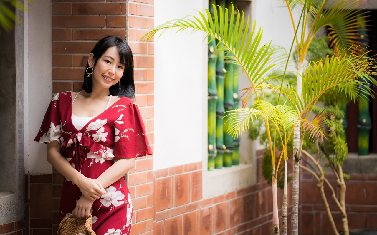 Улыбающаяся азиатка в красном платье стоит у стены 