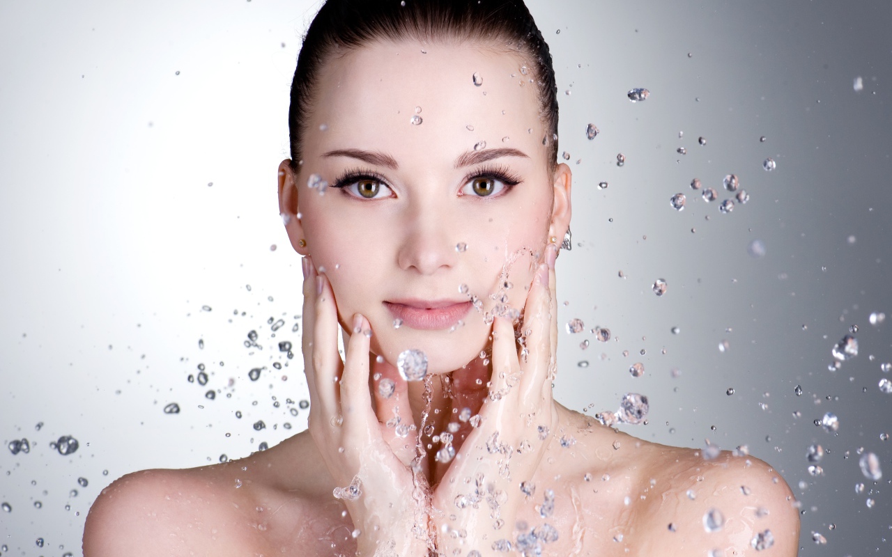 Брызги воды летят на красивое лицо молодой девушки на сером фоне