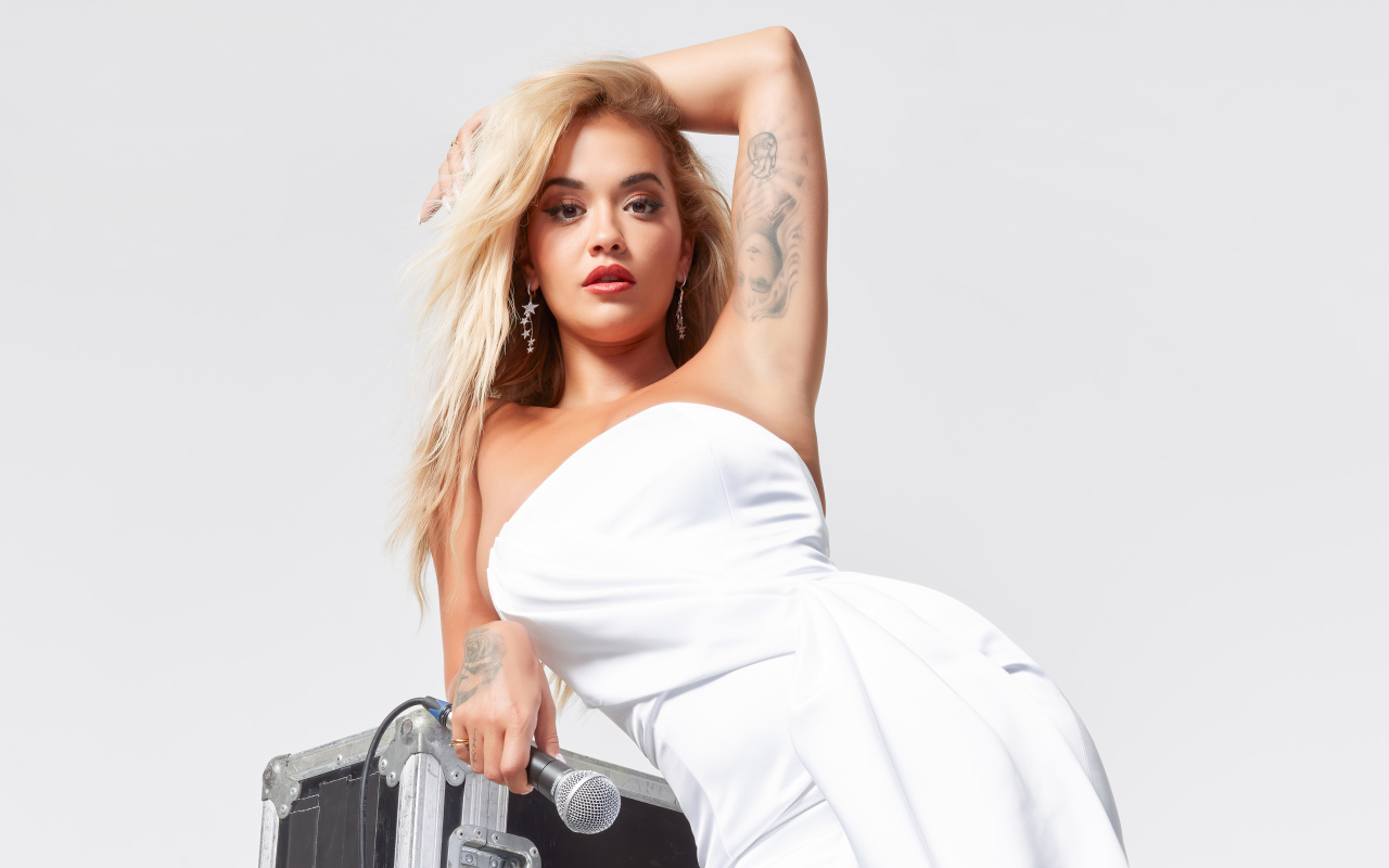 Певица Рита Ора в белом платье с микрофоном на сером фоне 