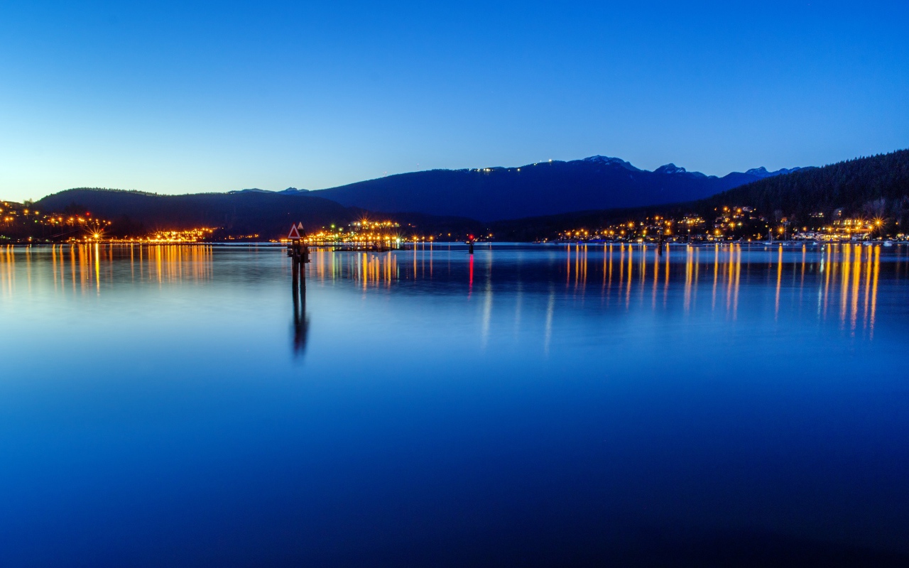 Огни ночного города отражаются в воде у гор 