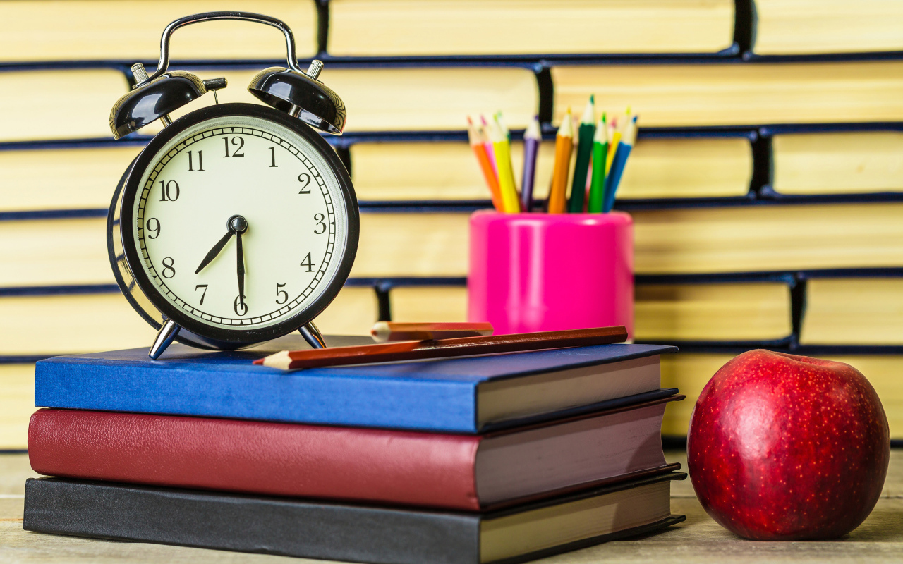 Будильник, книги, карандаши и яблоко на столе у стены 