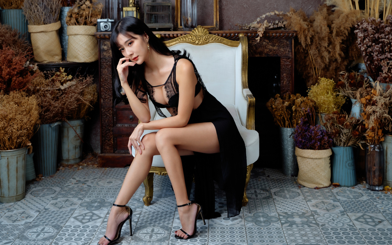 Красивая девушка азиатка в черном платье сидит в белом кресле 